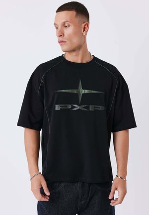 Man met een zwarte oversized t-shirt met een grote groene ster en "PXP"-logo, donkere jeans, kettingketting en oorbellen, kijkt naar links.