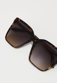 Tortoiseshell sunglasses with a large square frame, gradient dark lenses, and a glossy finish. Features subtle color variations in the frame.