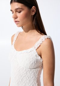 Bílá krajková top s volány na ramínkách, čtvercovým výstřihem, složitými květinovými vzory a jemnou texturou, předváděný na modelce.