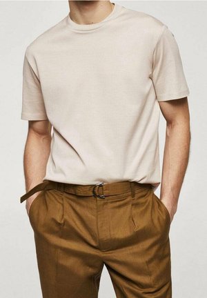 Hombre con camiseta beige claro de manga corta y lisa, metida dentro de pantalones marrones de talle alto con cinturón, con las manos en los bolsillos.