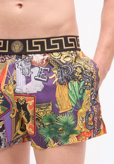 Έγχρωμα μαγιό βερμούδα με μωβ βάση, ζωντανά σχέδια, χρυσά μεταλλικά στοιχεία και μαύρη μέση στολισμένη με σχέδιο ελληνικού κλειδιού.