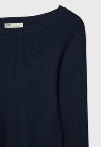 Pull en maille côtelée bleu marine avec col rond et manches longues, étiquette indiquant la taille et l'origine cousue à l'intérieur du col.
