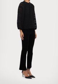 Donna che indossa una blusa nera trasparente e strutturata, pantaloni neri aderenti e decoltè neri con cinturino alla caviglia, in piedi su uno sfondo bianco.