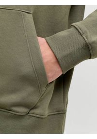 Tessuto di felpa verde oliva con tasca frontale, polsini a costine dalla texture ruvida e dettagli di cuciture visibili lungo i lati.