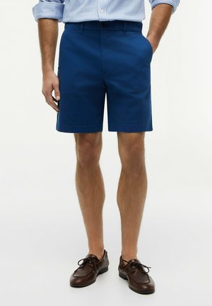 Man draagt marineblauwe korte broek, gestreept shirt met lange mouwen ingestopt, en donkerbruine leren veterschoenen, met één hand in de zak.