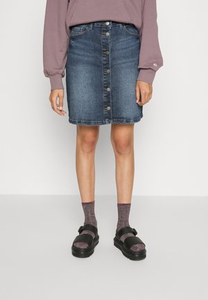 Person, die einen Jeansrock mit Knopfleiste, ein mauvefarbenes Langarmshirt, glitzernde violette Socken und schwarze Sandalen trägt und auf einem grauen Boden steht.