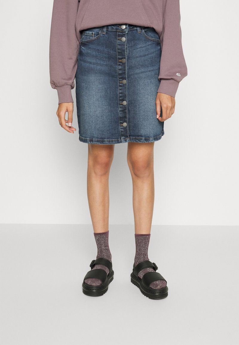 Personne portant une jupe en jean à boutons devant, un haut mauve à manches longues, des chaussettes violettes scintillantes et des sandales noires, debout sur un sol gris.