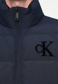 Námornícka prešívaná bunda s vysokým golierom, s textúrovaným čiernym vyšívaným logom „cK“ na prsiach, so zapínaním na zips a prešívaným dizajnom.