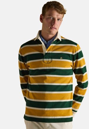 Man wearing a green, mustard yellow, and white striped long-sleeve rugby shirt with beige collar and blue undershirt, standing against a white background.