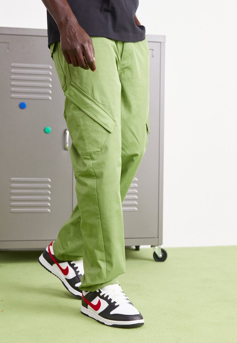 Persoon in lichtgroene cargo broek en zwart-witte-rode Nike sneakers, binnenshuis wandelend nabij een grijze locker op wielen.