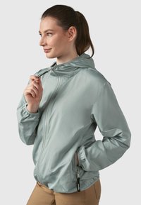 Gaiši zilā kapucēta jaka no gludas auduma, ar elastīgiem aprocēm, priekšējo rāvējslēdzēju un sānu kabatām ar rāvējslēdzēja aizdari.