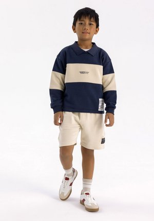 Felpa a righe blu navy e crema con colletto, abbinata a pantaloni beige e sneakers bianche con dettagli rossi e calze a coste.