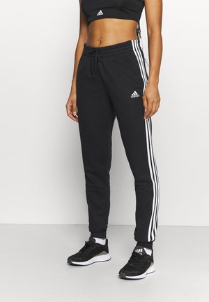 Femme portant un pantalon de survêtement Adidas noir avec des bandes blanches et des baskets noires, debout devant un fond blanc uni.