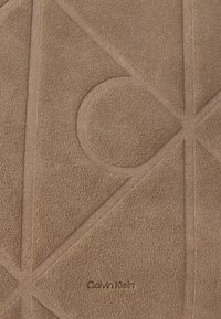 Közeli felvétel egy texturált barna felületről, amelyen kölcsönösen keresztbe álló domború vonalak geometrikus formákat alkotnak, valamint a "Calvin Klein" felirat domború textúrával. 