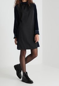 Frau in einem schwarzen Kleid mit samtigen langen Ärmeln, durchsichtigen schwarzen Strumpfhosen und schwarzen Leder-Schnürstiefeletten, die mit einem Bein übereinander steht.