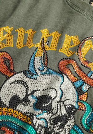 Oliwkowa koszula z błyszczącymi kryształkami. Projekt przedstawia czaszkę z kolorowymi akcentami i wyraźnym żółtym napisem. Teksturowany, codzienny materiał.