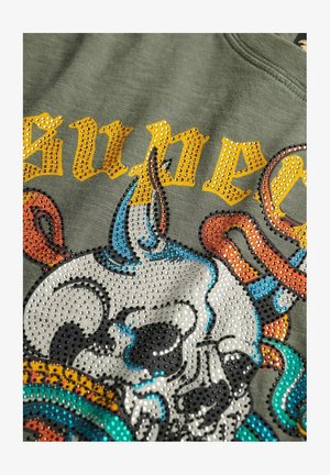 Olivengrøn skjorte med skinnende rhinsten. Designet inkluderer en kranium med farverige accenter og fed, gul tekst. Tekstureret, afslappet stof.