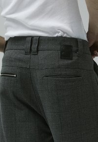 Gros plan d'une personne portant un pantalon gris à carreaux avec une ceinture noire et une chemise blanche côtelée rentrée, montrant les poches arrière et les passants de ceinture.
