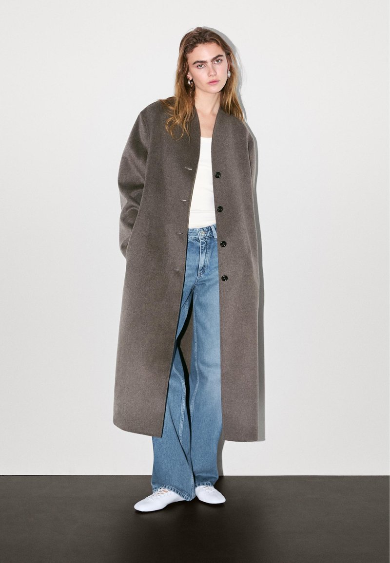 Langgrijze jas met een ontspannen pasvorm, voorzien van een ronde kraag en voorknopsluiting. Gecombineerd met lichtblauwe jeans en witte sneakers.