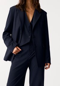 Costume bleu marine à fines rayures avec une veste ajustée et un pantalon à jambes larges. La veste comporte deux poches avant et trois boutons, la texture du tissu est lisse.