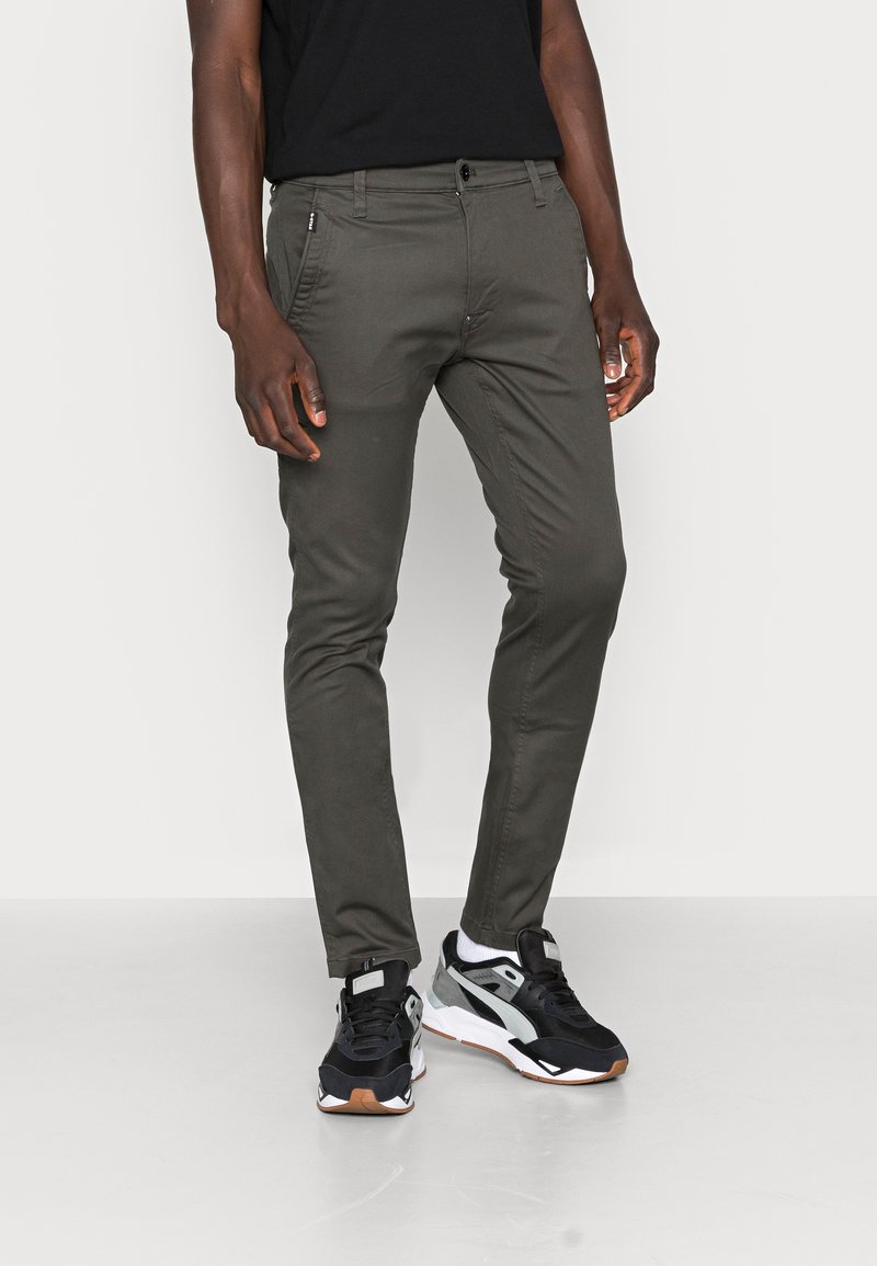 Dark green slim-fit trousers made of cotton blend material, featuring a zip pocket and button closure, paired with black and white sneakers.
