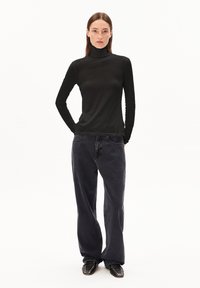 Zwarte langemouwen turtleneck top, gecombineerd met losse grijze jeans. Het model draagt zwarte gespijkerde schoenen, wat zorgt voor een contrasterende textuur en stijl.