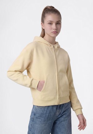 Giovane donna con i capelli biondi che indossa una felpa con zip giallo chiaro e jeans blu, in piedi con una mano in tasca.