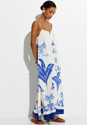 Woman wearing a white maxi dress with blue tropical leaf prints, thin straps, and brown flat sandals, standing against a white background.
