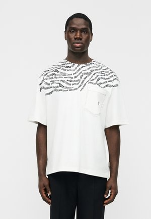 Mężczyzna noszący białą, oversize'ową koszulkę z powtarzającym się czarnym napisem „Just Cavalli” na ramionach i kieszeni na piersi, stojący na jednolitym tle.