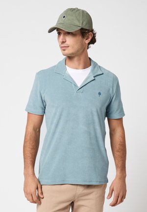 Homme portant un polo à manches courtes bleu clair, un maillot blanc en dessous, un pantalon beige et une casquette olive avec un logo d'arbre, regardant vers la gauche.