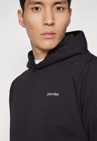 Černá mikina s hladkým materiálem. Obsahuje kapuci se stahovací šňůrkou a malé bílé logo "Calvin Klein" na hrudi. Detailní pohled.
