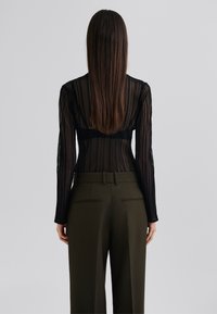 Svart blonde bodysuit med lange ermer, med vertikale striper og gjennomsiktig tekstur, kombinert med olivengrønne høytlivs bukser.