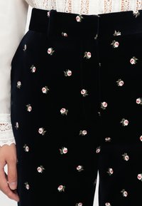 Detailní záběr na černé sametové kalhoty s drobně vyšívanými růžovými a bílými květinami, které jsou nošeny s bílou halenkou s krajkovými manžetami.