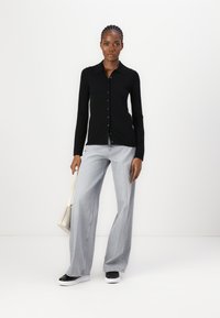 Camicia nera a maniche lunghe con bottoni, jeans grigi a gamba larga e sneakers nere; tiene in mano una piccola borsa bianco sporco. Design minimalista con una vestibilità classica.