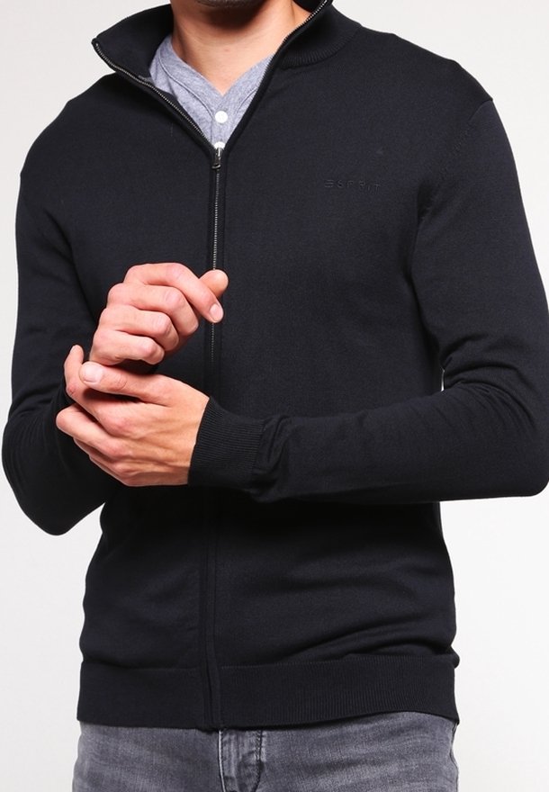 Svart zippered tröja i stickad material med hög krage och ribbade ärmslut, som har en subtil logotyp på bröstet, bärs över en grå skjorta.