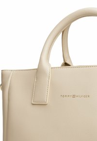 Borsa in pelle beige con doppio manico superiore, superficie testurizzata e logo Tommy Hilfiger impresso. Presenta bordi cuciti e un design minimalista.