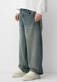 Suurjalad denim püksid heledas sinise-halli värvitoonis, kontrastsete õmblustega ja ees olevate taskutega, kombineeritud heledate tossudega.