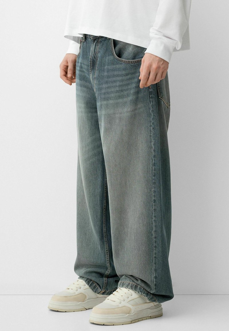 Suurjalad denim püksid heledas sinise-halli värvitoonis, kontrastsete õmblustega ja ees olevate taskutega, kombineeritud heledate tossudega.