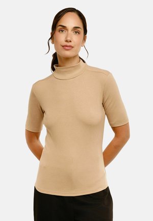 Kvinde med mørkt hår bundet tilbage iført en beige kortærmet rullekravetrøje og sorte bukser mod en ensfarvet lys baggrund.