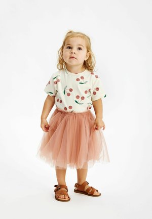 Un enfant en bas âge portant une chemise blanche avec des imprimés de fleurs rouges, une jupe en tulle rose, et des sandales marron à brides. Design de tenue simple et ludique.