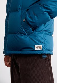 Veste matelassée bleue avec une texture lisse et brillante, design isolant, et étiquette de marque à l'ourlet. Associée à un pantalon en velours côtelé marron.