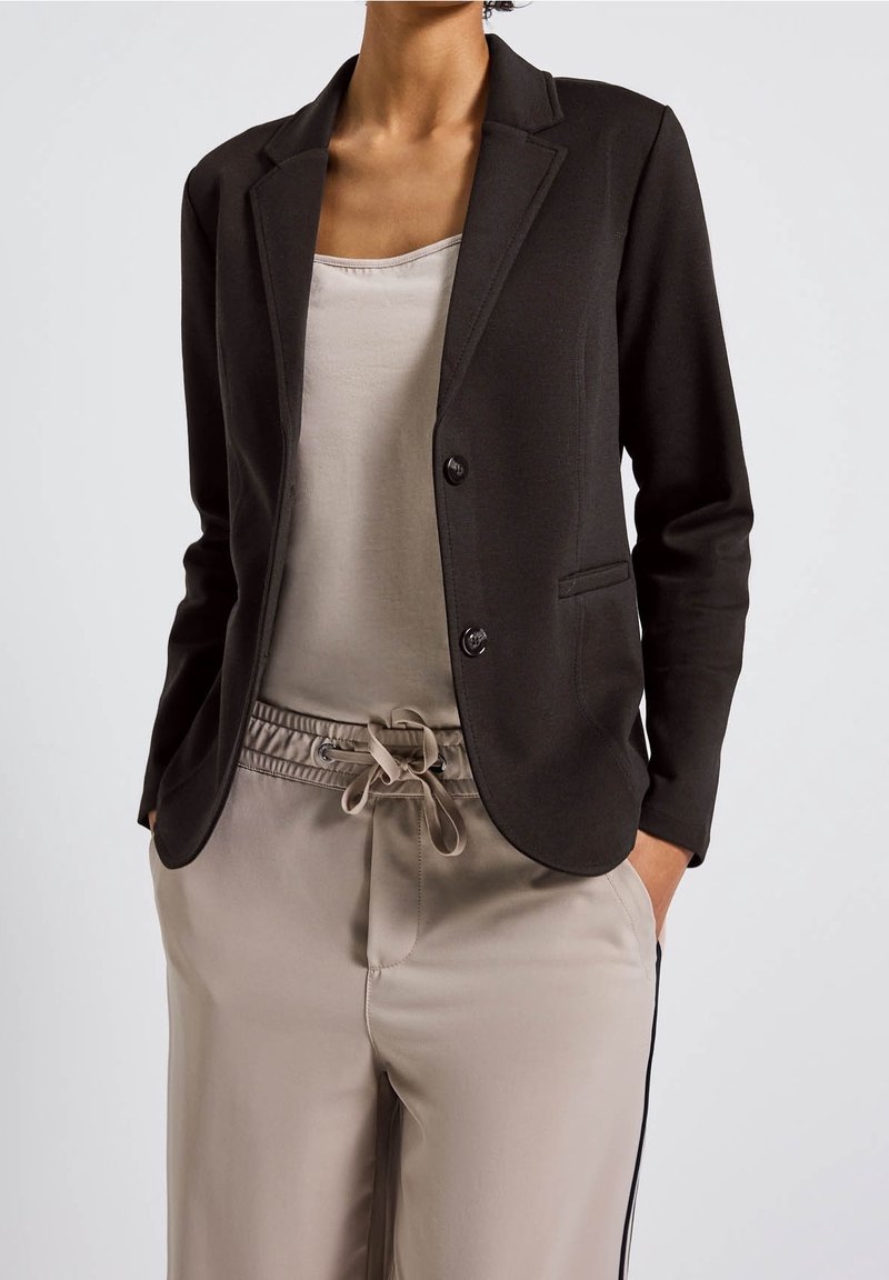 Personne portant un blazer noir sur un haut beige clair et un pantalon taupe à cordon, mains dans les poches, vue des épaules aux cuisses.