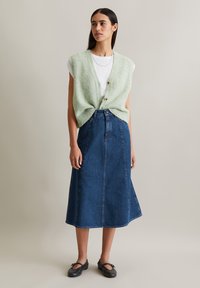 Gilet sans manches en tricot vert avec des boutons sur un t-shirt blanc, associé à une jupe en denim longue. Des chaussures noires à enfiler complètent la tenue.