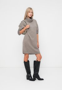 Vestido de malha bege com gola alta e mangas curtas, combinado com botas pretas de pele com salto bloco até ao joelho. Acessório com uma mala de couro com padrão de cobra.