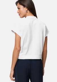 Haut court à manches courtes blanc avec un col, présentant une texture lisse et un design épuré, associé à un pantalon bleu marine foncé.