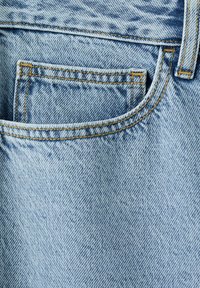 Jean en denim bleu clair avec une petite poche avant pour pièces de monnaie et des coutures jaunes visibles autour des bords de la poche.