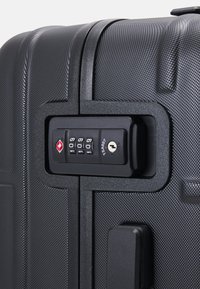 Crna tvrda kofer s teksturiranom površinom, opremljena kombinacijskim bravom i TSA odobrenim ključem, osigurana crnim trakama.