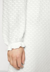 Vestido branco com tecido texturizado apresentando um padrão sutil, mangas longas com punhos franzidos, exibindo uma aparência delicada e suave.