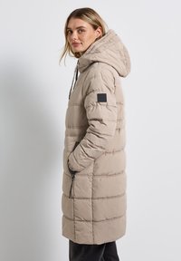 Beige prošívaný kabát s kapucí, vybavený kapsou na zip a texturovanými panely. Hlavní materiál má hladký povrch a moderní, prodlouženou siluetu.