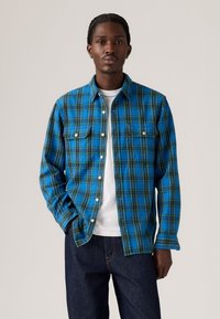 Blauw geblokt flanellen shirt met groene en gele lijnen, twee borstzakken en witte knopen, gedragen over een wit t-shirt.